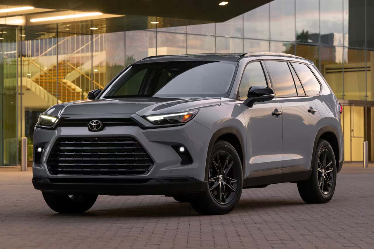 Gray 2025 Toyota Grand Highlander Hybrid parked in front of a modern glass building, showcasing its sleek profile, LED headlights, and upscale SUV styling.