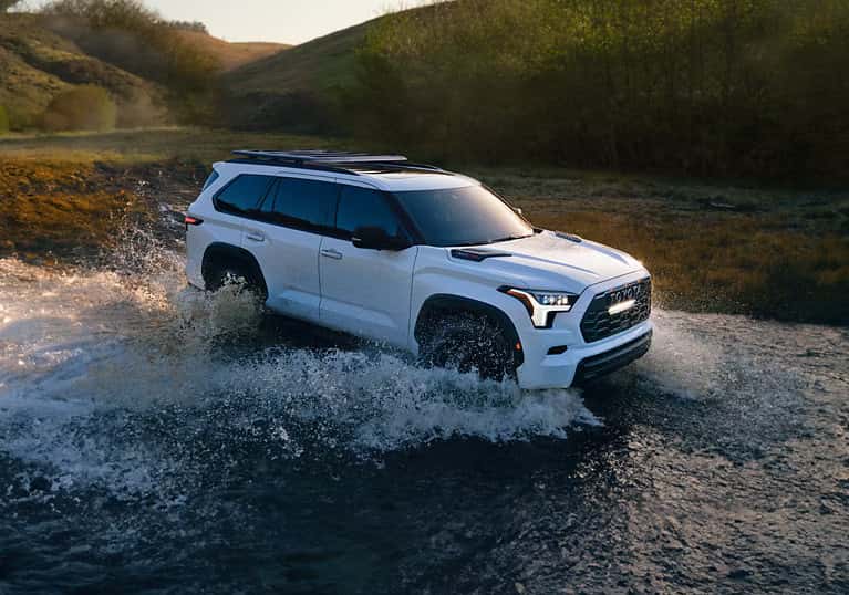 White 2025 Toyota Sequoia driving through a shallow stream with water splashing, showcasing off-road capability against a scenic, hilly background at sunset.