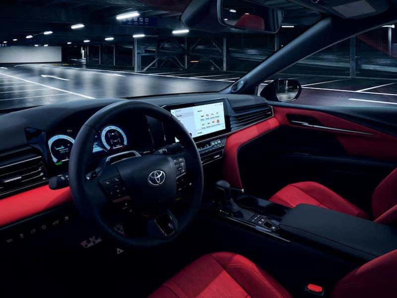 The front seats and dashboard of a 2026 Toyota Camry sedan, featuring red upholstery accents and a touchscreen display, with a dark parking structure visible past the windshield
