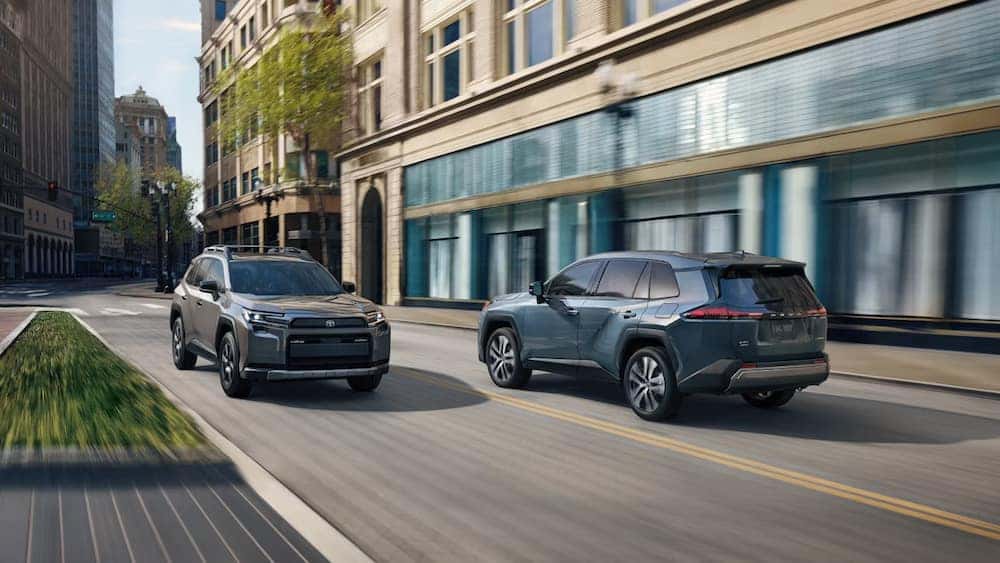 Two 2026 Toyota RAV4 SUVs, one gray and one blue, driving in opposite directions down a city street