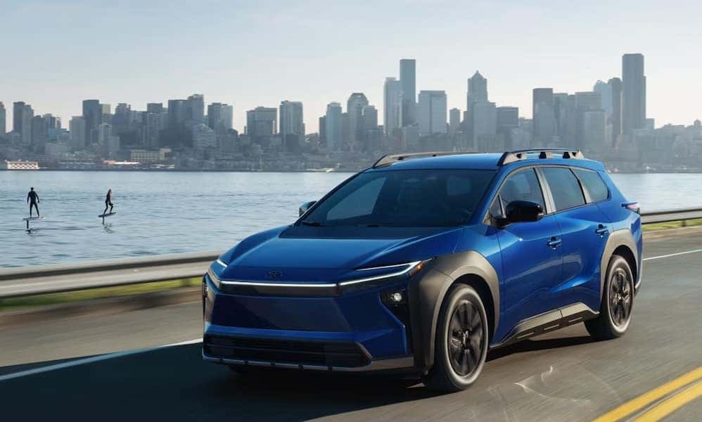 A bright blue 2026 Toyota bZ Woodland SUVdriving down a road with a body of water and a city skyline in the distance
