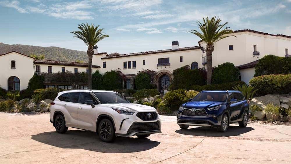 Two 2026 Toyota Highlander SUVs, one white and one blue, parked in front of a large house