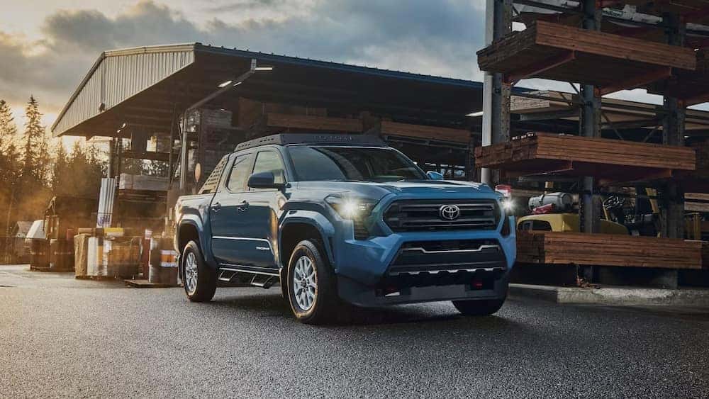 A blue 2026 Toyota Tacoma pickup truck parked outside a hardware store with its headlights on