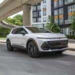 Front 3/4 view of a 2024 Chevrolet Equinox EV RS in Summit White driving in a downtown area.