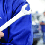 A cropped image of a Honda technician's shoulder and hand holding a wrench. His blue polo shirt shows the Honda logo, and the blurred background shows a Honda service center.