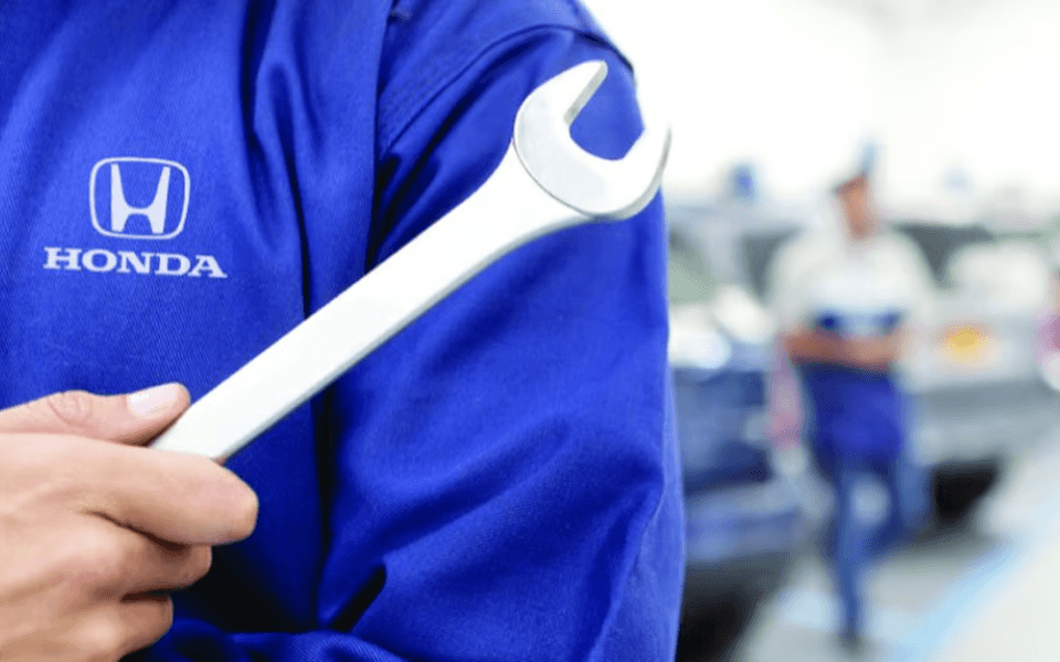A cropped image of a Honda technician's shoulder and hand holding a wrench. His blue polo shirt shows the Honda logo, and the blurred background shows a Honda service center.