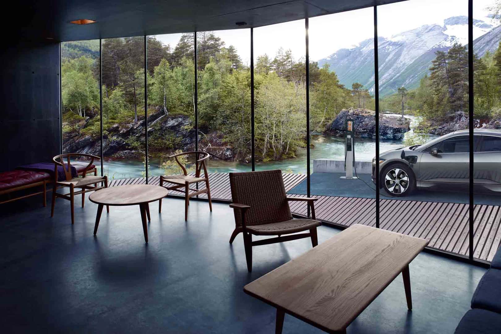 Jaguar I-PACE charging outside a glass-walled cabin by a scenic river and mountains