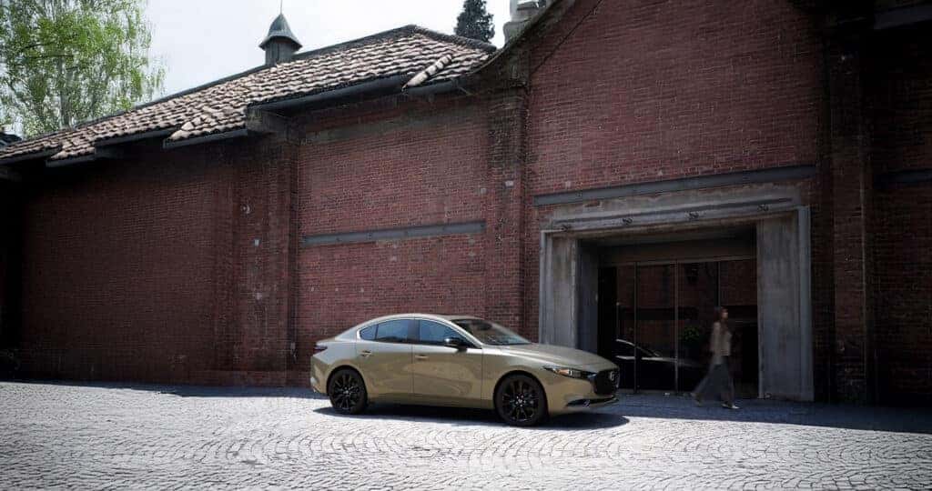 2025-Mazda3-Zicron-Sand-parked-in-front-of-the-store-front-with-a-women-walking-by