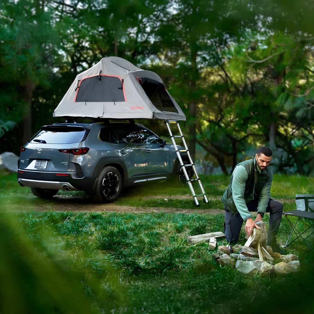 2025 Mazda CX-50 with a rooftop tent in the woods with a man building a campfire in the evening