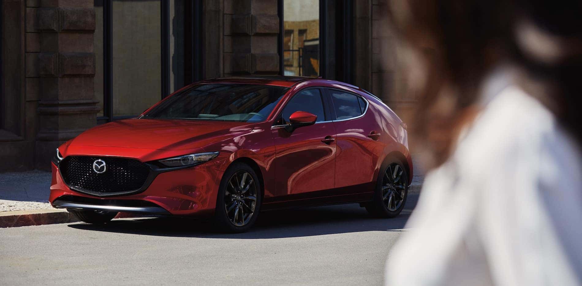 2026 Mazda3 hatchback in red parked on a stree with a figure of a woman looking at it