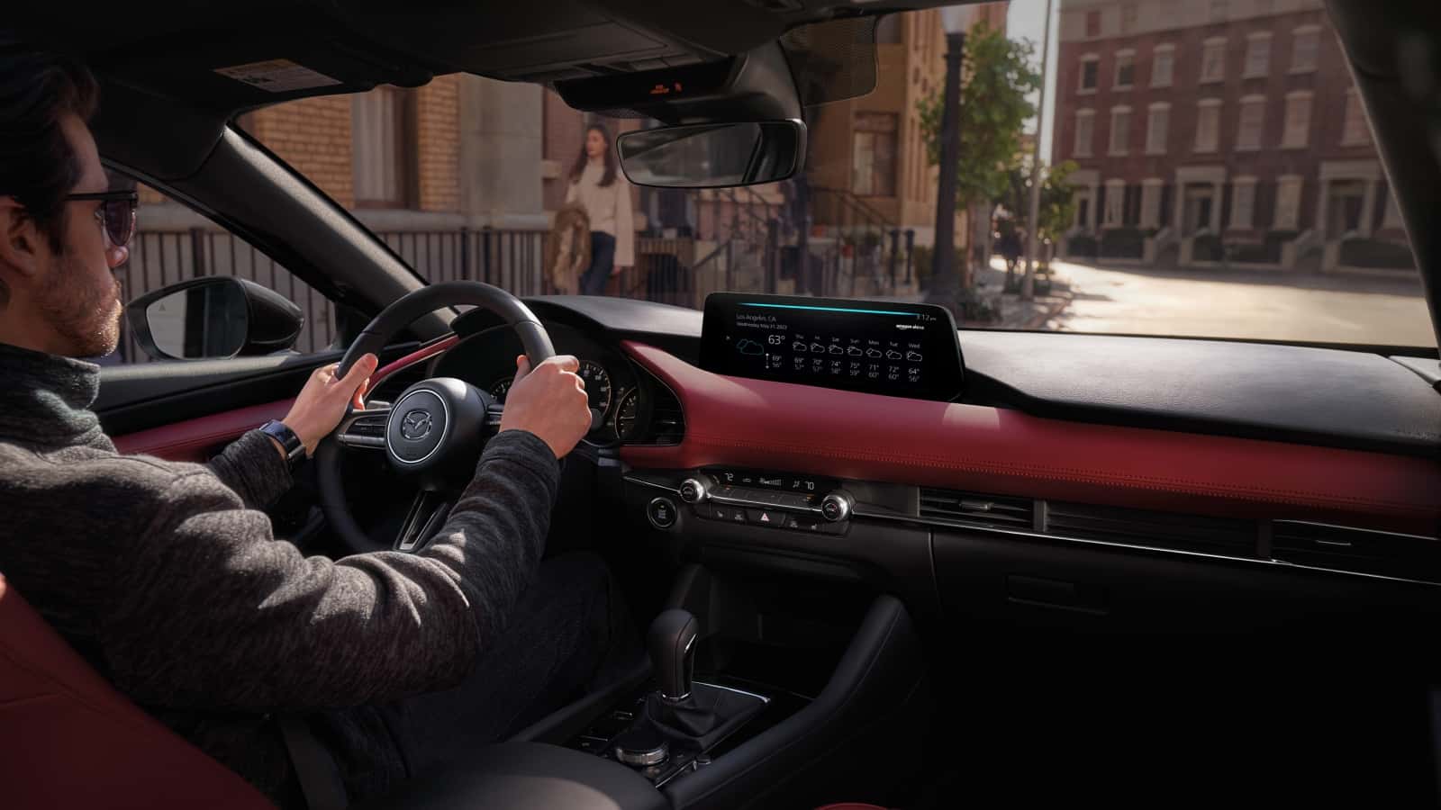 2026 Mazda3 interior view from the front seats showing the dashboard, center display, red-trim cabin, and driver perspective in an urban setting with natural daylight.
