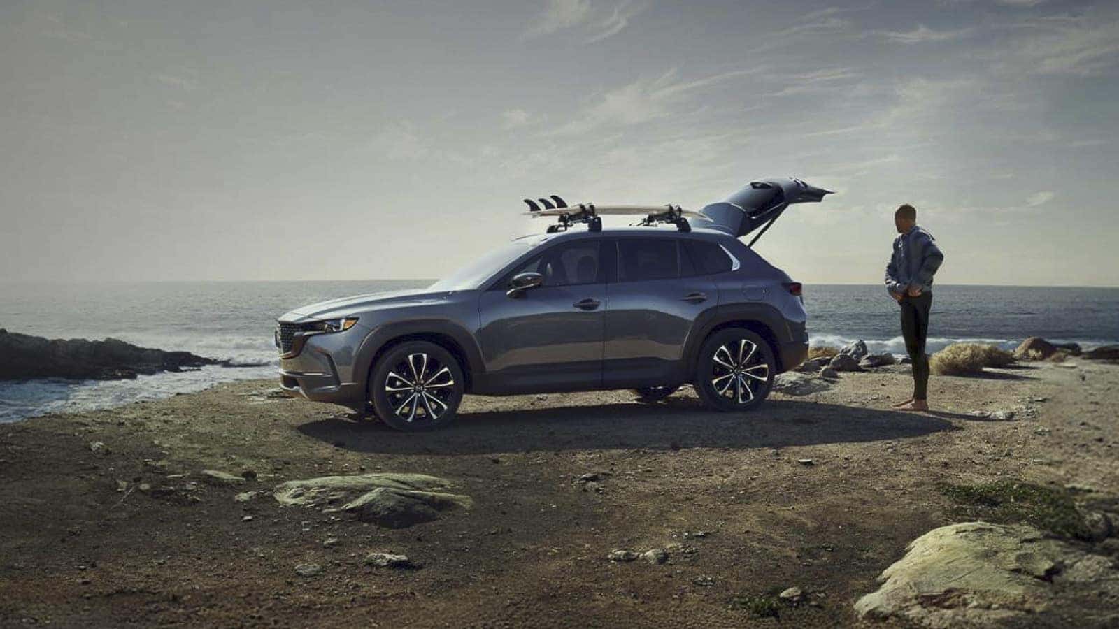 2026 Mazda CX-50 parked along a rocky coastline with ocean views, rear hatch open and roof rack carrying outdoor gear