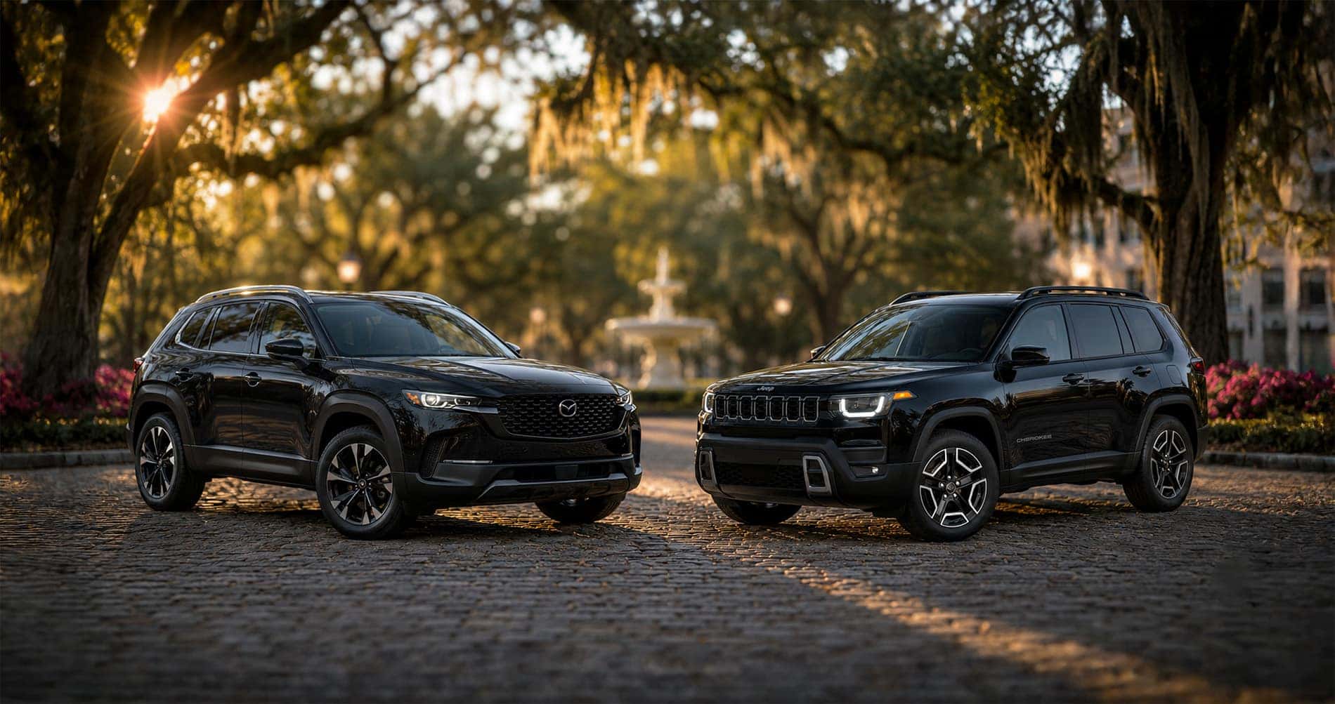 Black Mazda CX-50 Hybrid and black Jeep Cherokee Hybrid parked on a cobblestone street near a fountain at sunset, emphasizing styling and proportions.