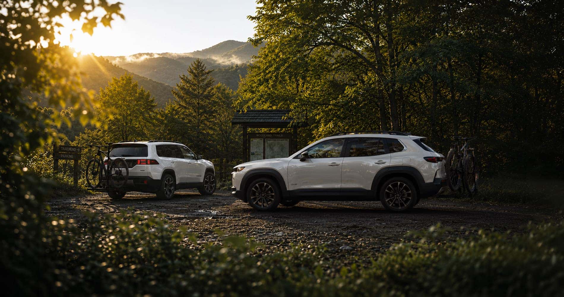 White Mazda CX-50 Hybrid and white Jeep Cherokee Hybrid parked near a trailhead with bicycles mounted, highlighting outdoor utility and rear design.