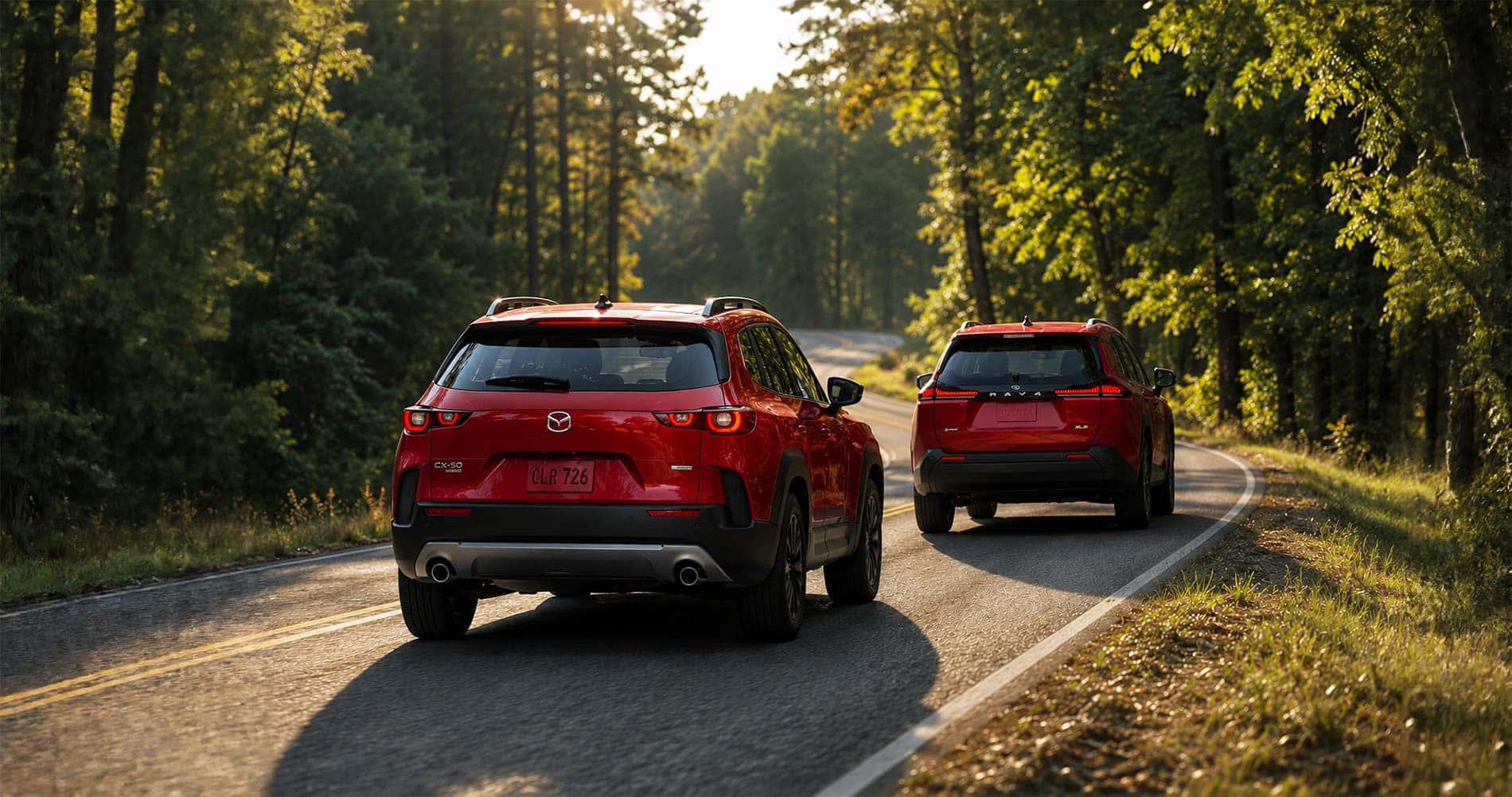 Red Mazda CX‑50 Hybrid and red Toyota RAV4 Hybrid driving on forest road at sunset highlighting rear design and taillight styling.