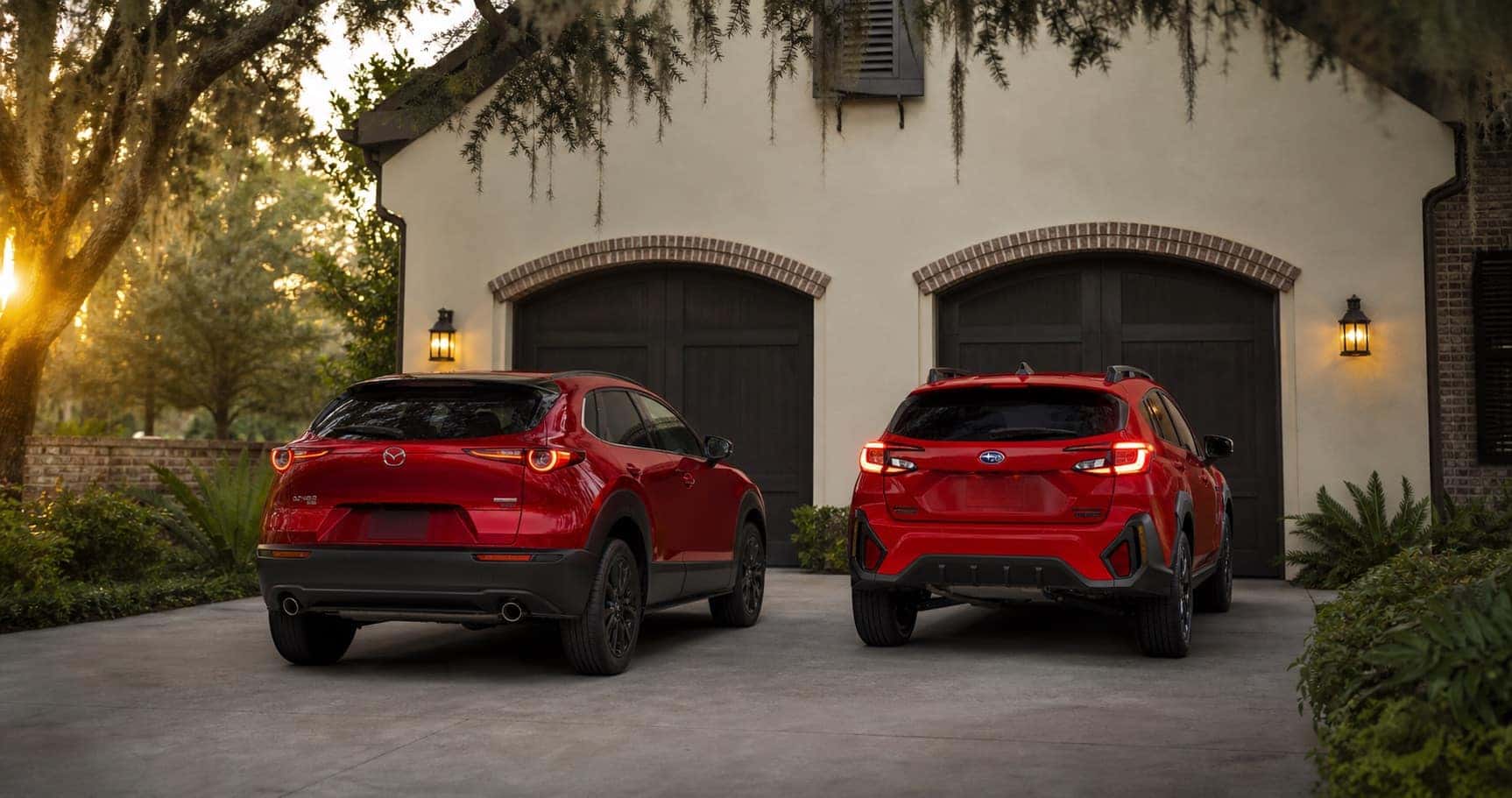 Red Mazda CX‑30 and red Subaru Crosstrek parked side by side in driveway at sunset in front of brick home highlighting rear styling and design differences.