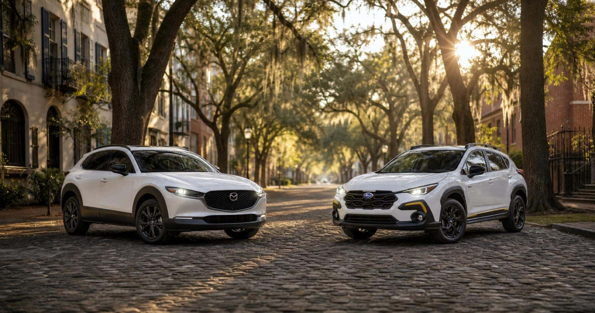 White Mazda CX‑30 and white Subaru Crosstrek parked on cobblestone street under tree canopy at sunrise showcasing front grille designs and trim level styling.