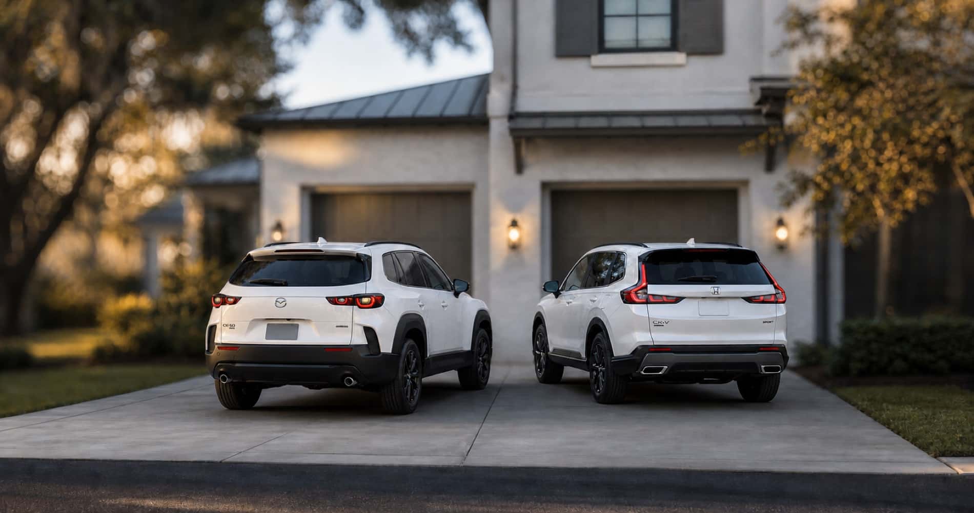 White Mazda CX‑50 Hybrid and white Honda CR‑V Hybrid parked side by side in driveway at sunset in front of brick home highlighting rear styling and hybrid badging differences.