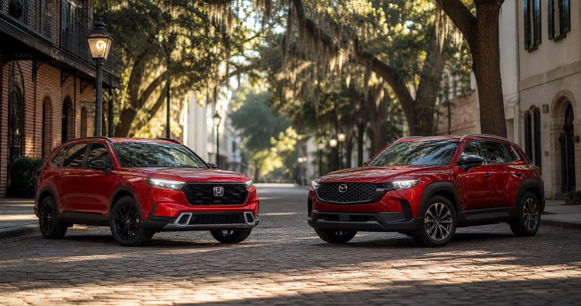 Red Mazda CX‑50 Hybrid and red Honda CR‑V Hybrid parked on cobblestone street under tree canopy at sunrise showcasing front grille designs and trim level styling.