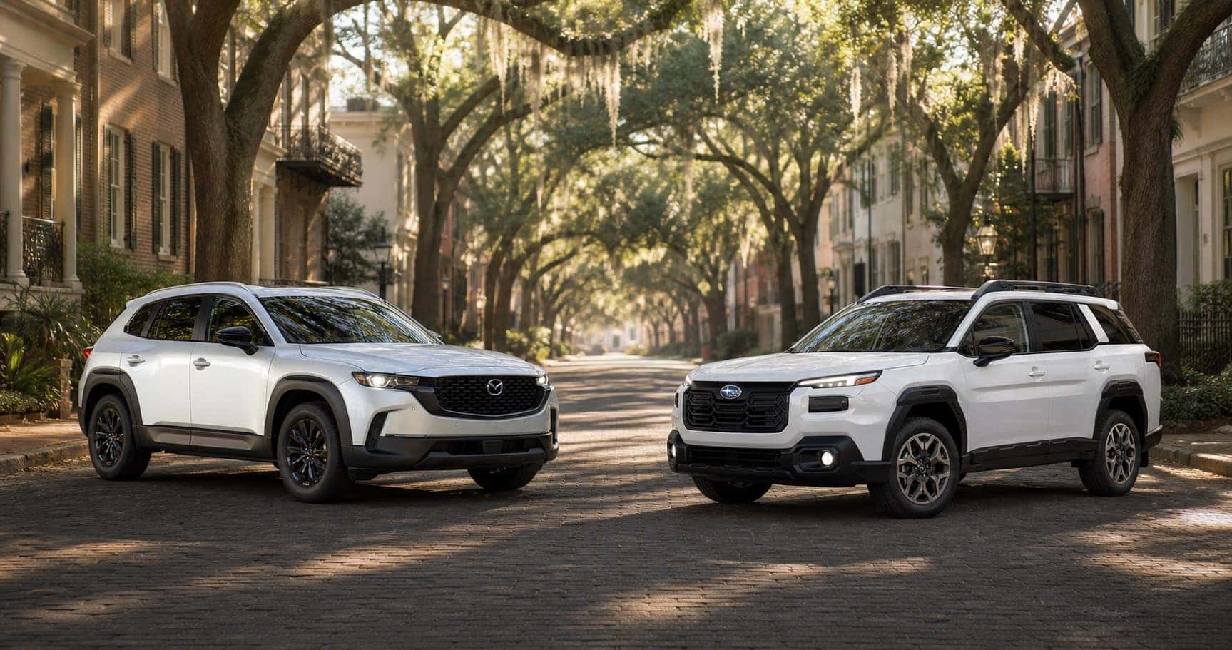 White Mazda CX‑50 and white Subaru Outback parked on cobblestone street under tree canopy at sunrise showcasing front grille designs and trim level styling.