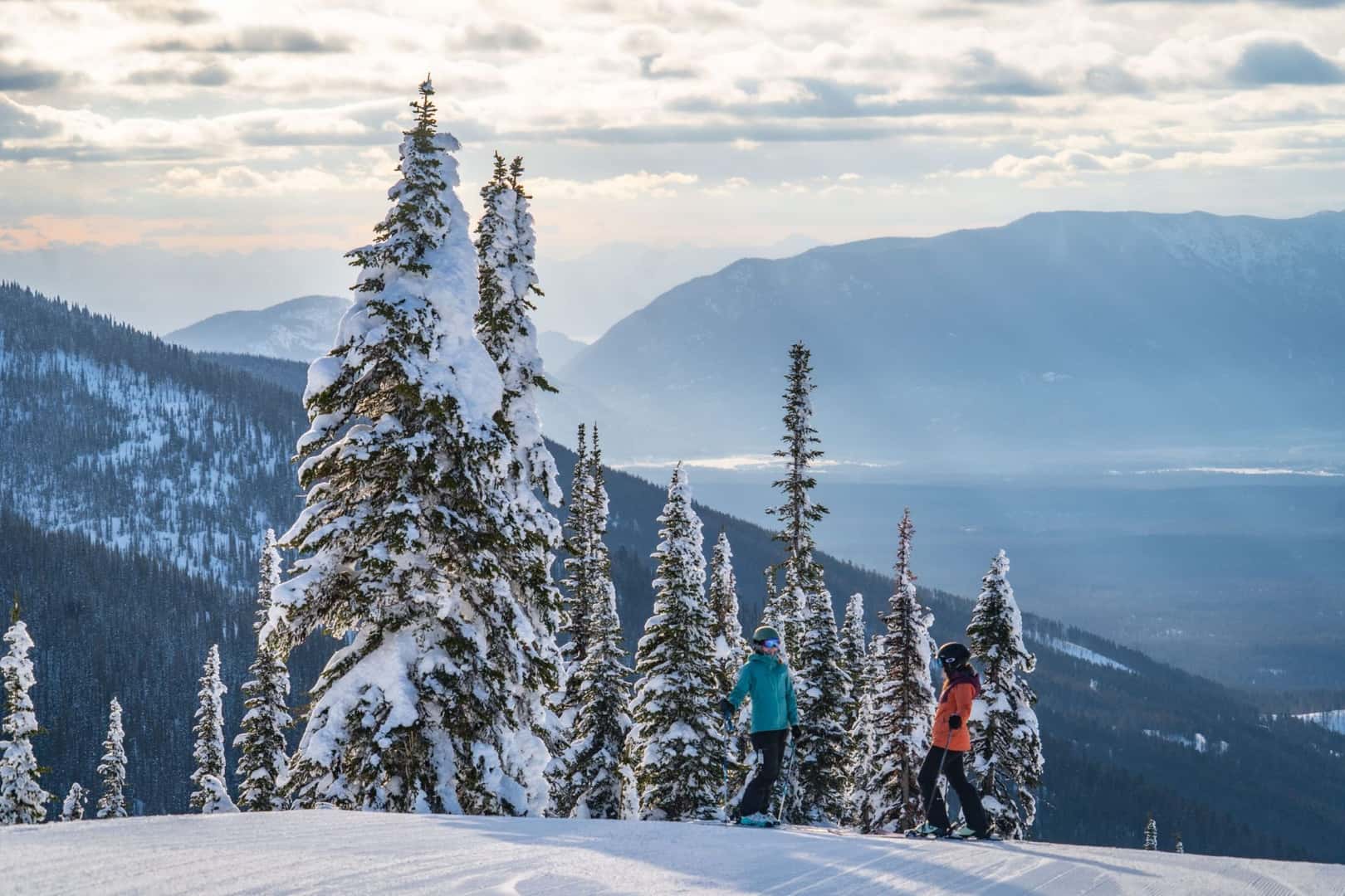 Two skiers stand on top of a snowy mountain overlooking the Flathead Valley