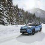 Blue Toyota Rav4 drives on a snowy road with trees and mountains in the background