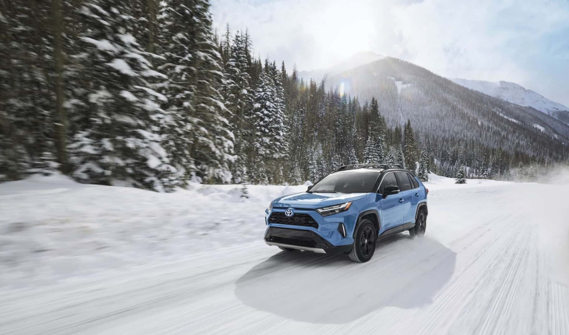 Blue Toyota Rav4 drives on a snowy road with trees and mountains in the background