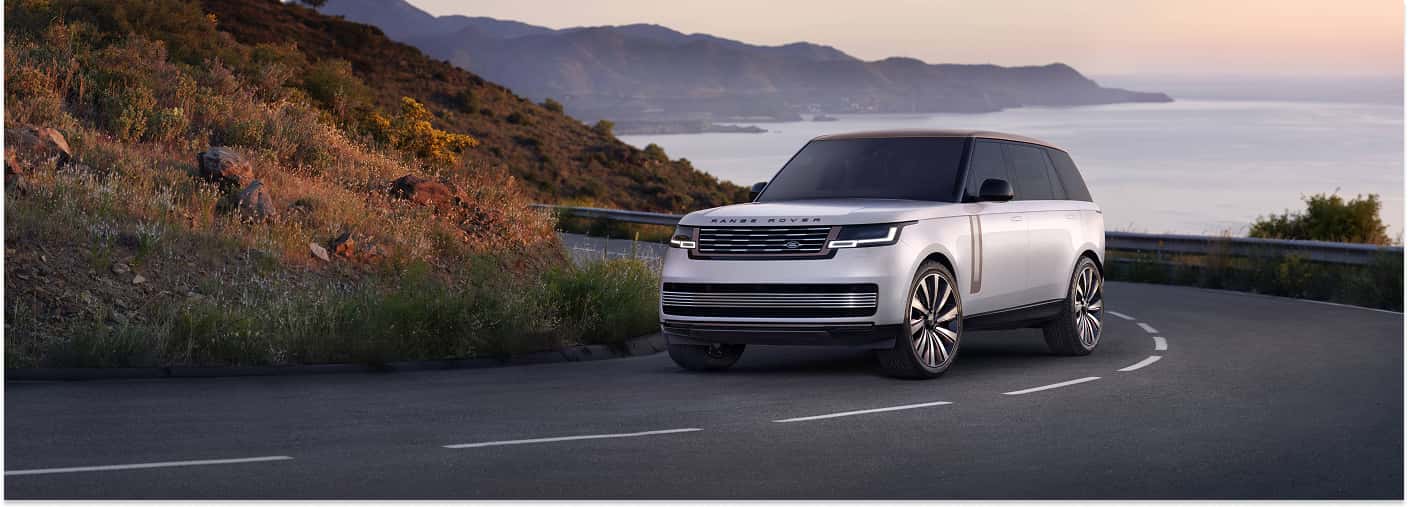 A white 2025 Range Rover with a contrasting bronze roof is positioned on a curved asphalt road overlooking the ocean and coastal mountains at sunset.