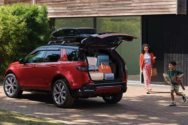 A red Land Rover SUV with the tailgate open and luggage loaded inside, as a woman and child walk toward it carrying bags