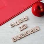 Scrabble pieces spelling out "Back to School" on a wooden desk with red folder and apple.