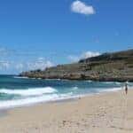View of the beach from afar with waves crashing on the shore and a hill in the background