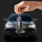 A hand is shown holding a car key in front of a blurred black sedan.