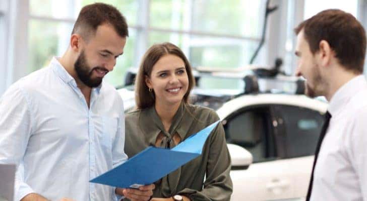 A couple is shown speaking to a car salesman at a car dealership.