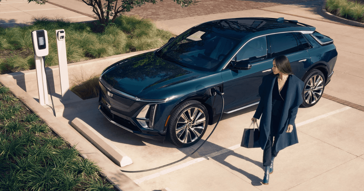 A lighter blue 2025 Cadillac LYRIQ parked and plugged in at a charging station as a woman walks away from it