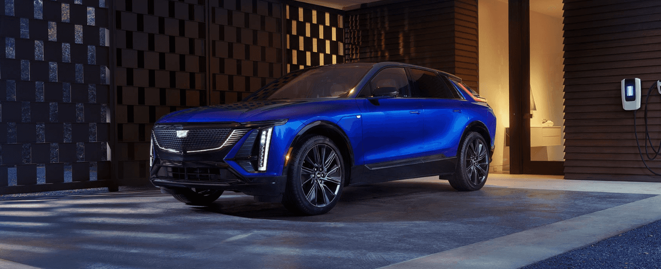 A blue Cadillac LYRIQ parked in front of a house by a charging station