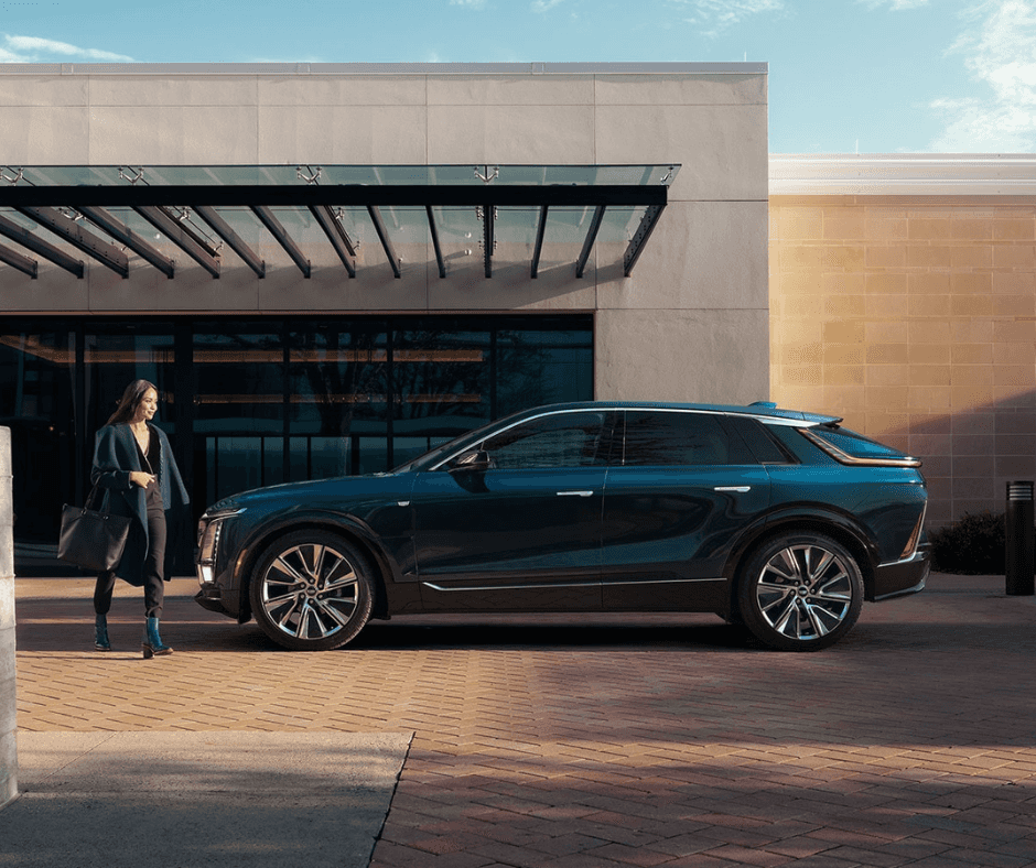 A blue Cadillac LYRIQ in profile, parked in front of a building as a woman walks in front of it