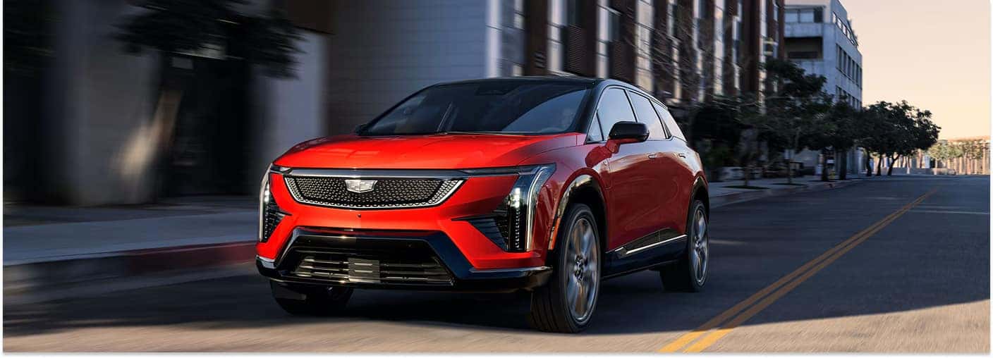 **Option 1:** Red 2026 Cadillac OPTIQ driving on an urban street, shown from a front three-quarter angle.  **Option 2:** Front view of a red 2026 Cadillac OPTIQ in motion on a city street with buildings in the background.
