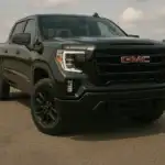 Front angle view of a black GMC Elevation pickup truck parked on an open lot under a partly cloudy sky, showcasing its bold grille, black wheels, and modern headlight design.