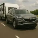 A gray Buick Enclave driving on a highway while towing a large white travel trailer, illustrating the vehicle’s towing capability on an open road under a partly cloudy sky.